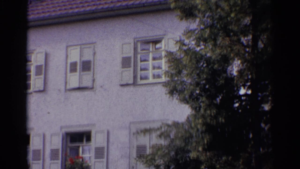 1961 Germany 8mm frame of a shuttered stucco building with a tiled roof edge and leafy branches partly obscuring the facade.