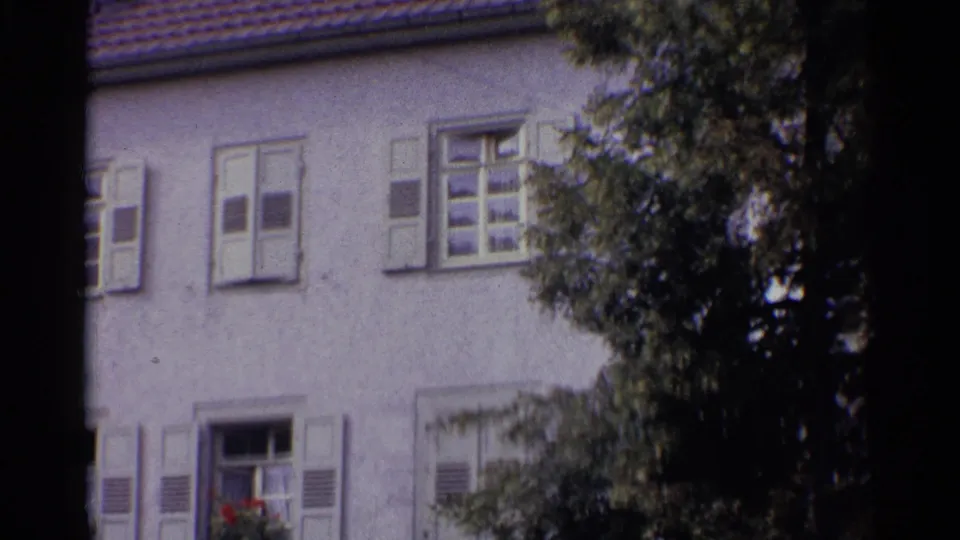 1961 Germany 8mm frame of a shuttered stucco building with a tiled roof edge and leafy branches partly obscuring the facade.