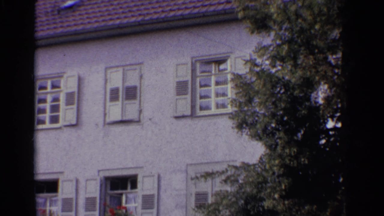 Pale stucco house facade with shuttered windows and red roof tiles partly framed by trees.