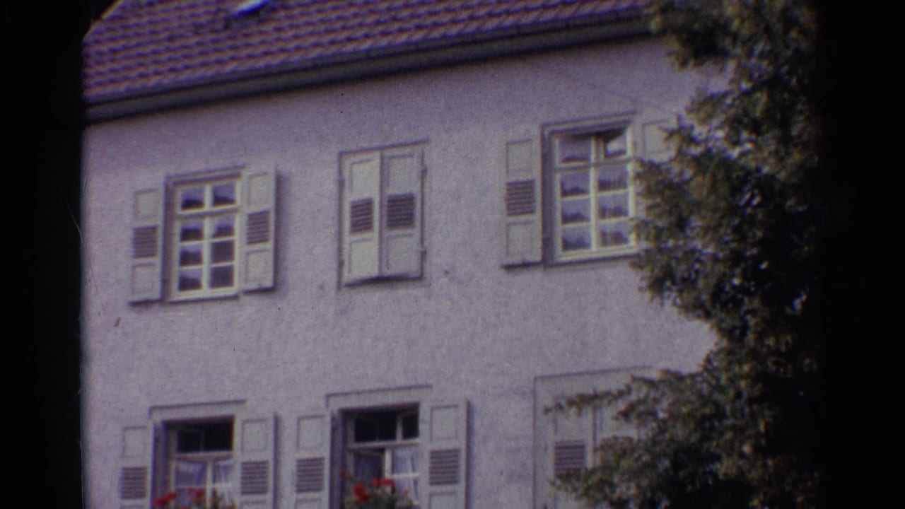 Two-story stucco house with shuttered windows, roof tiles, and leafy branches at the right edge.