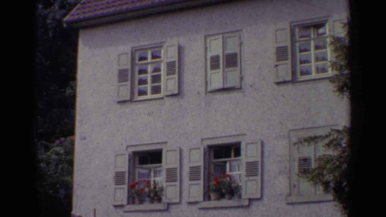 1961 Germany frame showing multiple shuttered windows on a pale stucco residence, with red flower boxes and leafy foliage at the edge of the shot.