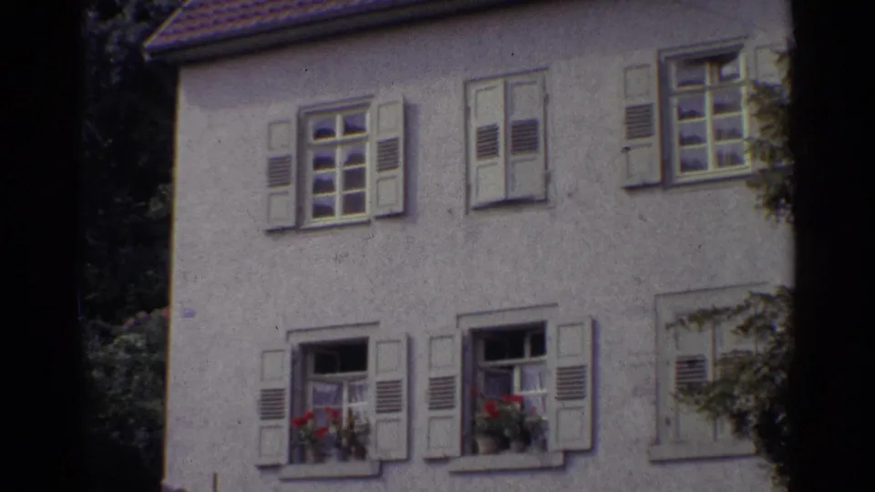 1961 Germany frame showing multiple shuttered windows on a pale stucco residence, with red flower boxes and leafy foliage at the edge of the shot.