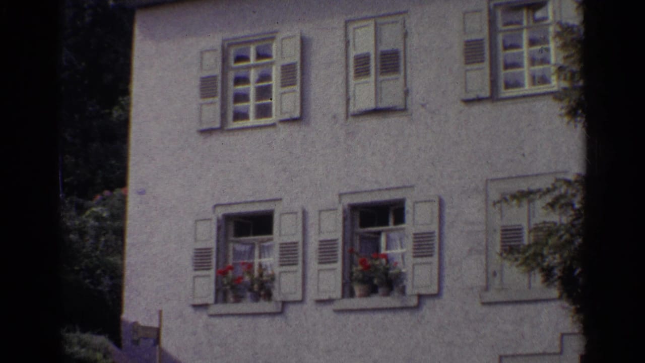 1961 Germany home-movie frame showing a white stucco residential facade with shutters, open windows, and red flower boxes.