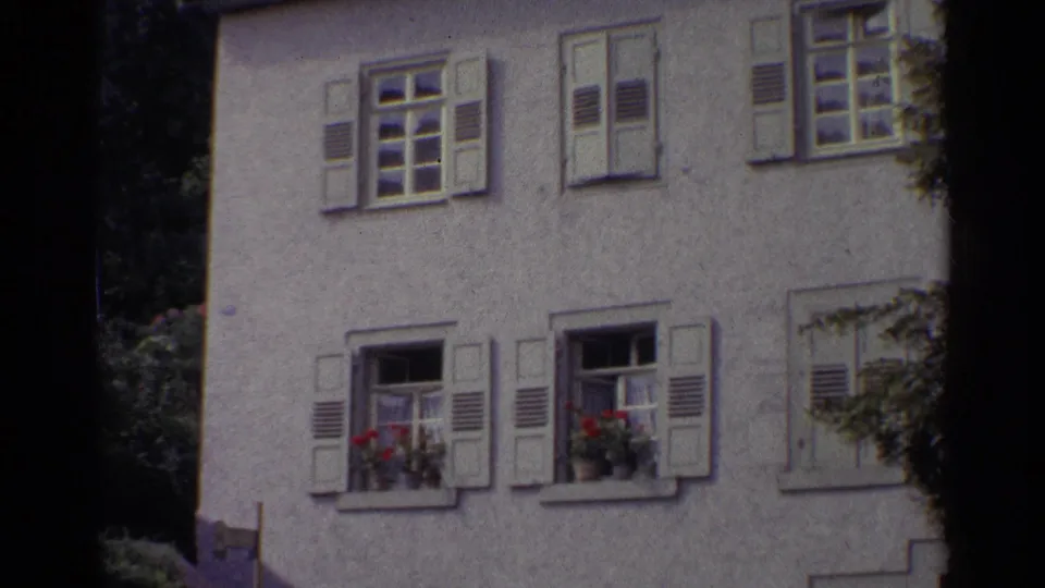 1961 Germany home-movie frame showing a white stucco residential facade with shutters, open windows, and red flower boxes.