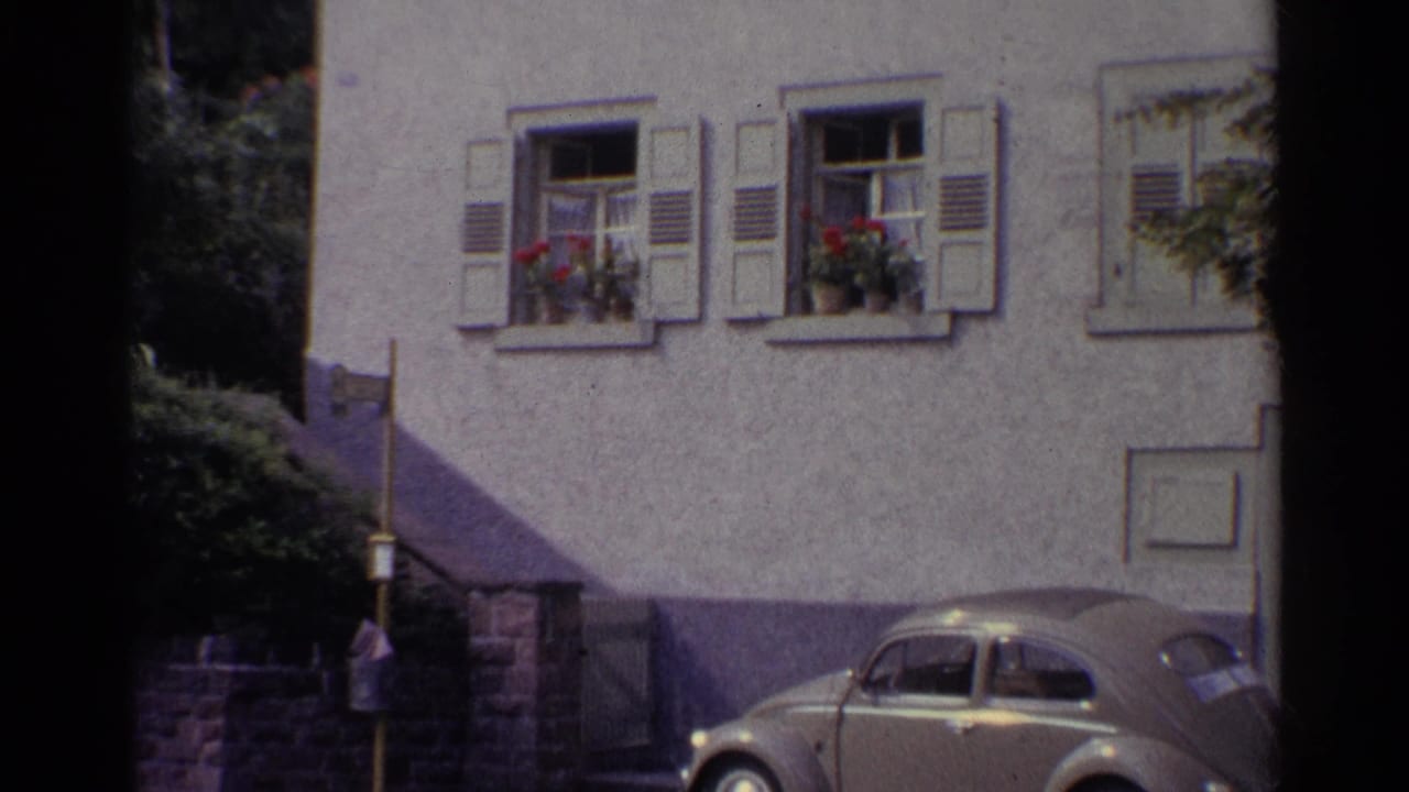 1961 Germany home-movie view of the lower facade, shuttered windows, flower boxes, stair rail, shrubs, and a parked rounded car.