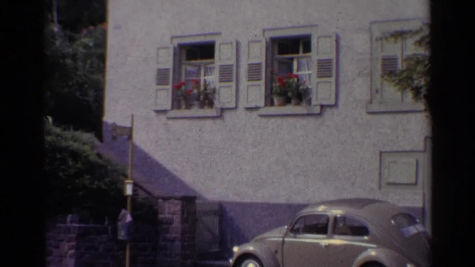 1961 Germany home-movie view of the lower facade, shuttered windows, flower boxes, stair rail, shrubs, and a parked rounded car.