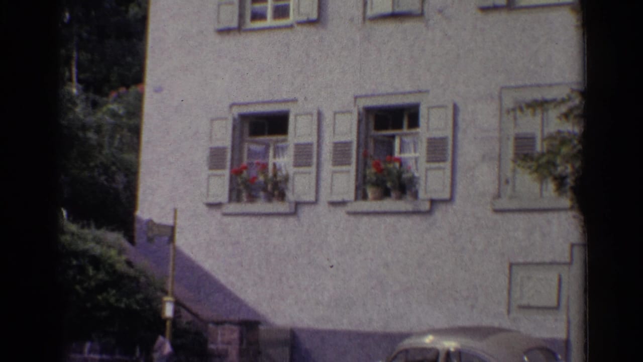 1961 Germany frame showing the white stucco building, shuttered windows, red flower boxes, shrubs, and a parked car near the base.