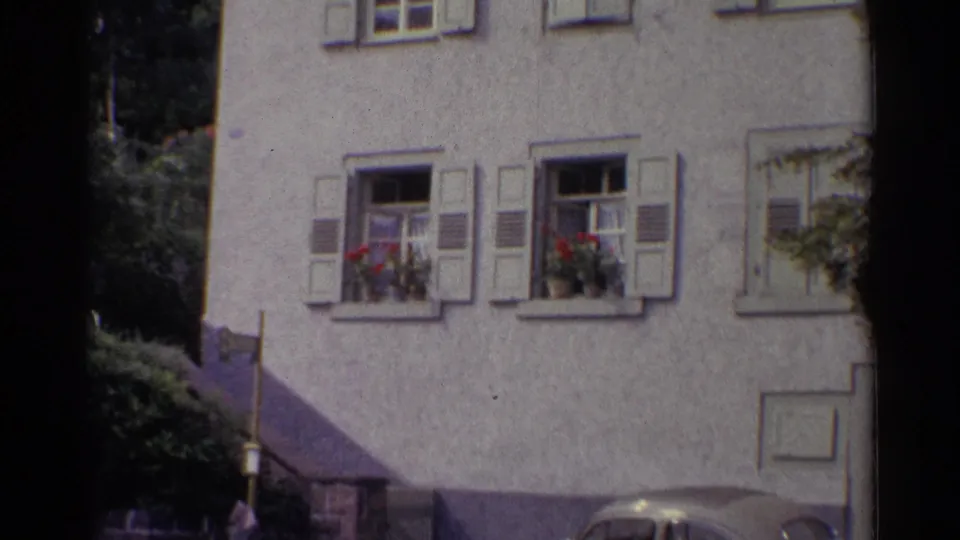 1961 Germany frame showing the white stucco building, shuttered windows, red flower boxes, shrubs, and a parked car near the base.