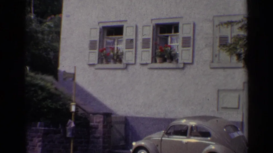 1961 Germany home-movie frame showing a Volkswagen Beetle parked along a quiet street below shuttered windows with red flowers.