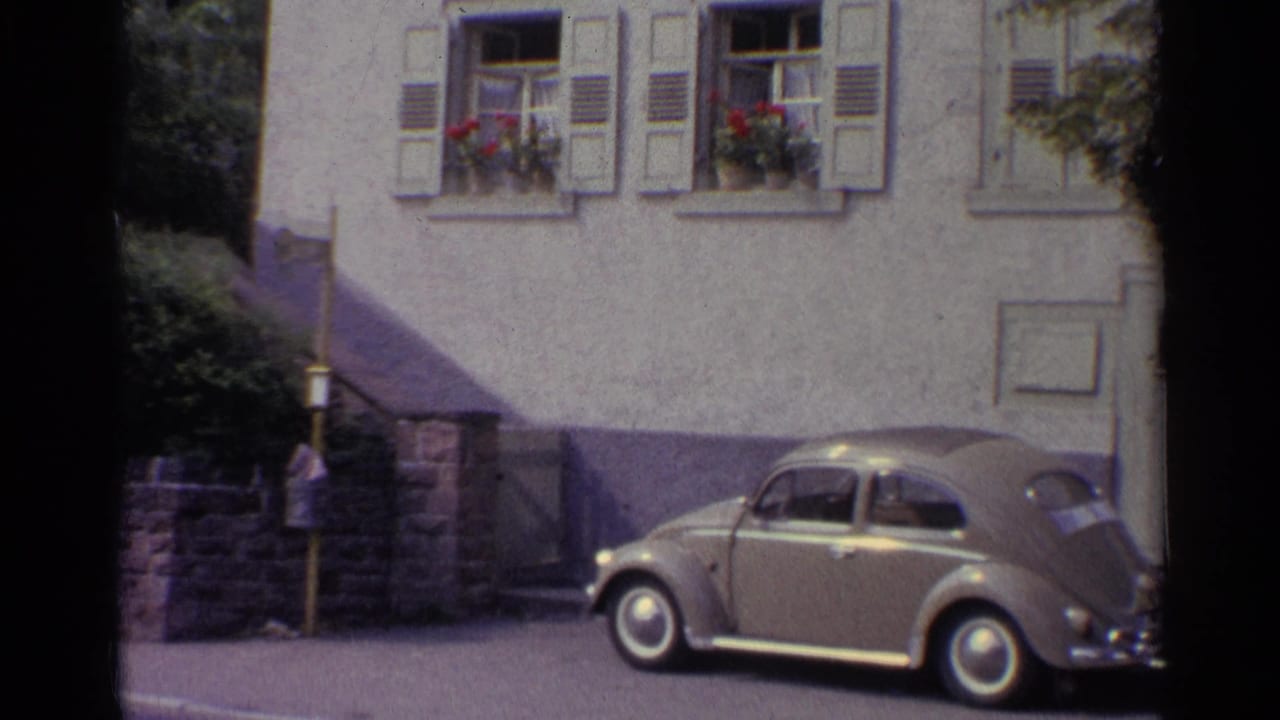 German 1961 street view with a parked Volkswagen Beetle, pale stucco wall, window shutters, flower boxes, and partial shadow across the building.