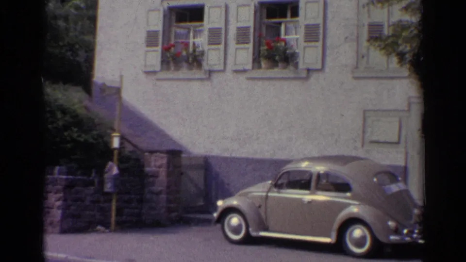 German 1961 street view with a parked Volkswagen Beetle, pale stucco wall, window shutters, flower boxes, and partial shadow across the building.