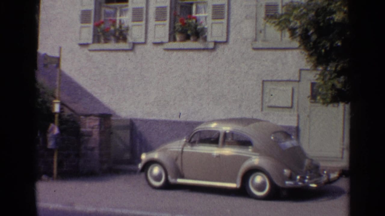 1961 Germany footage frame showing a parked Beetle near a doorway, shuttered windows, flower boxes, and a quiet town sidewalk.