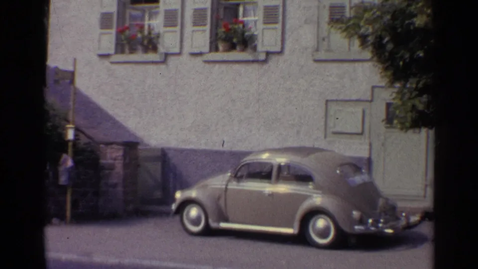 1961 Germany footage frame showing a parked Beetle near a doorway, shuttered windows, flower boxes, and a quiet town sidewalk.