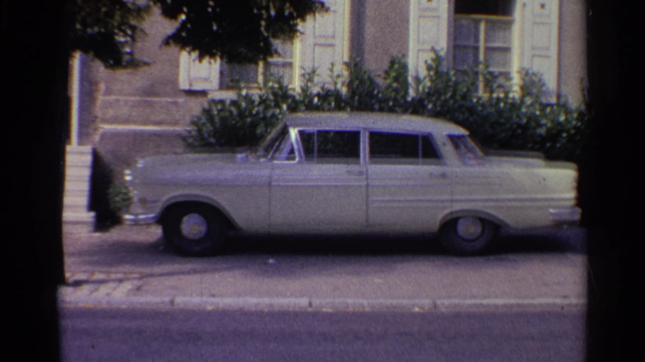 1961 Germany home-movie frame showing a larger classic sedan parked beside steps and shrubs in front of a shuttered building.