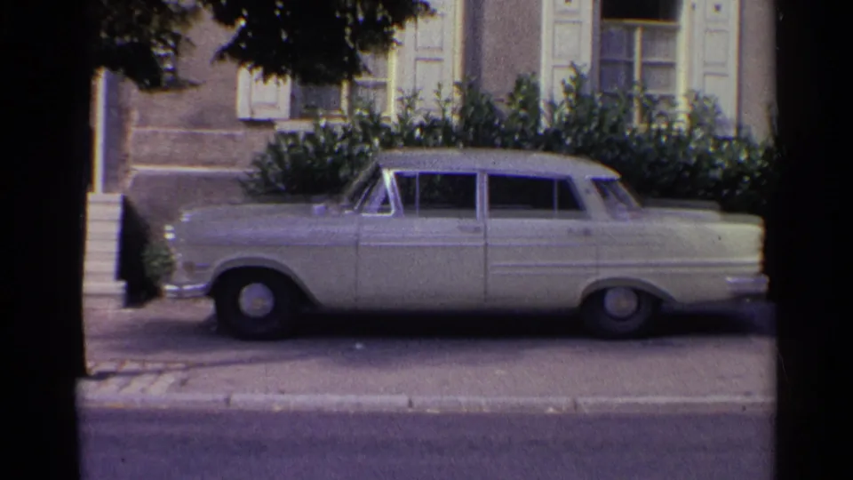 1961 Germany home-movie frame showing a larger classic sedan parked beside steps and shrubs in front of a shuttered building.