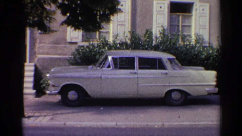 1961 Germany 8mm frame showing a white sedan parked along a residential street outside a shuttered house.
