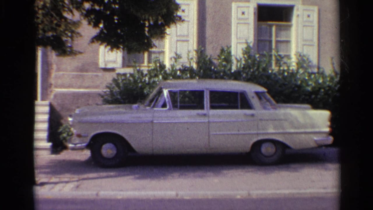 Parked white sedan in front of a house with pale shutters and garden shrubs.