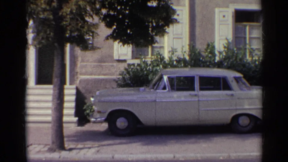 8mm view of a parked white sedan, front steps, tree trunk and shuttered house exterior in 1961 Germany.