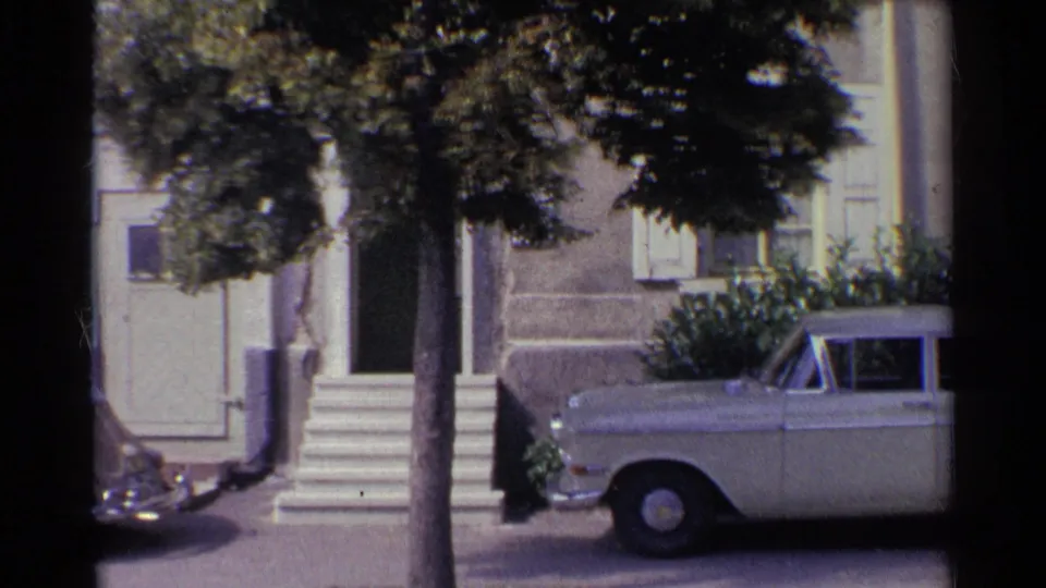 1961 Germany home-movie frame showing a pale sedan at the curb before a stucco house with white shutters, steps, and a street tree.