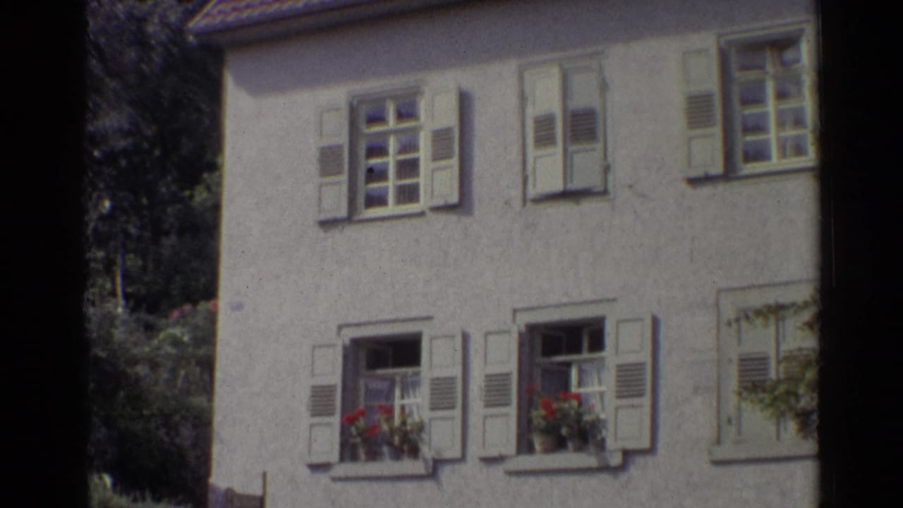 1961 Germany frame centered on a stucco house with white shutters, open lower windows, flower boxes, tiled roof, and surrounding trees.