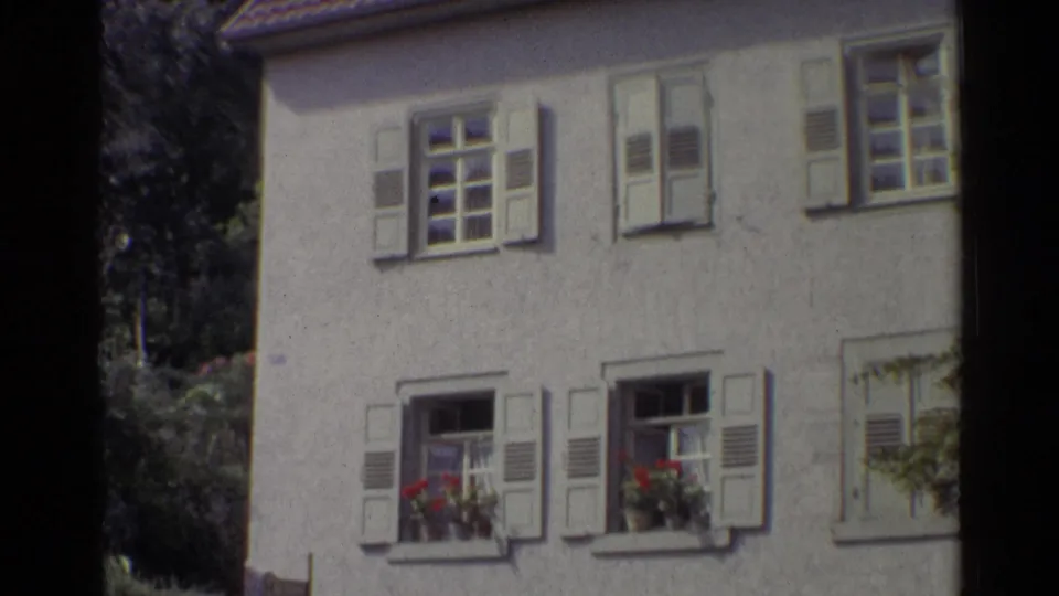 1961 Germany frame centered on a stucco house with white shutters, open lower windows, flower boxes, tiled roof, and surrounding trees.