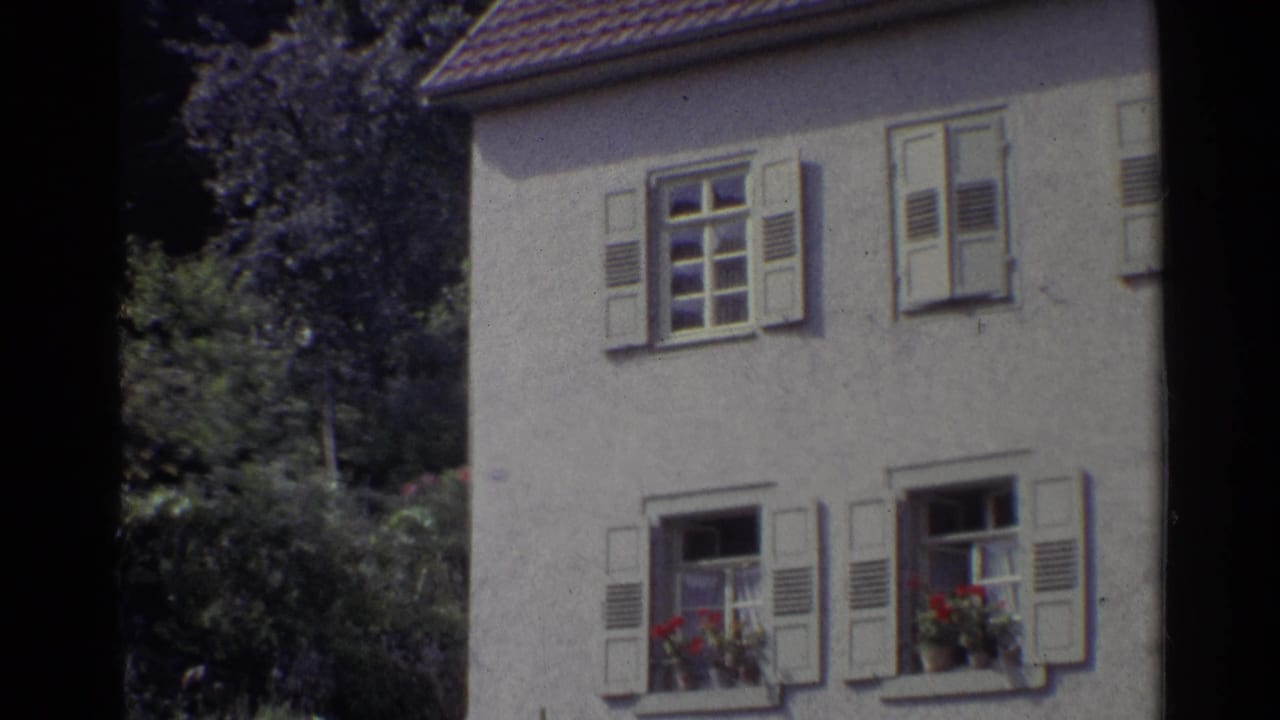 1961 Germany home-movie frame showing the upper and lower windows of a stucco house, with white shutters, flower boxes, roof tiles, and shrubs.
