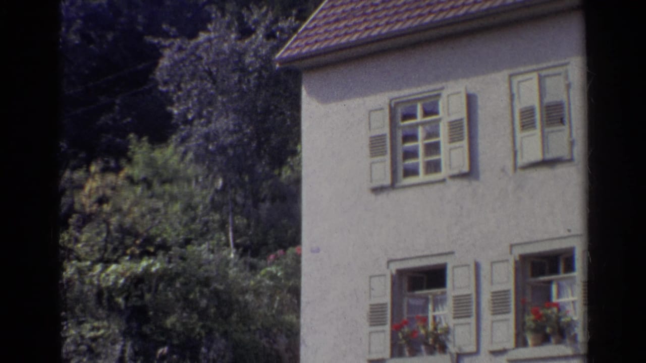 1961 Germany frame showing a shuttered stucco house at the edge of the composition, with flower boxes, hedges, trees, and a tiled roof.