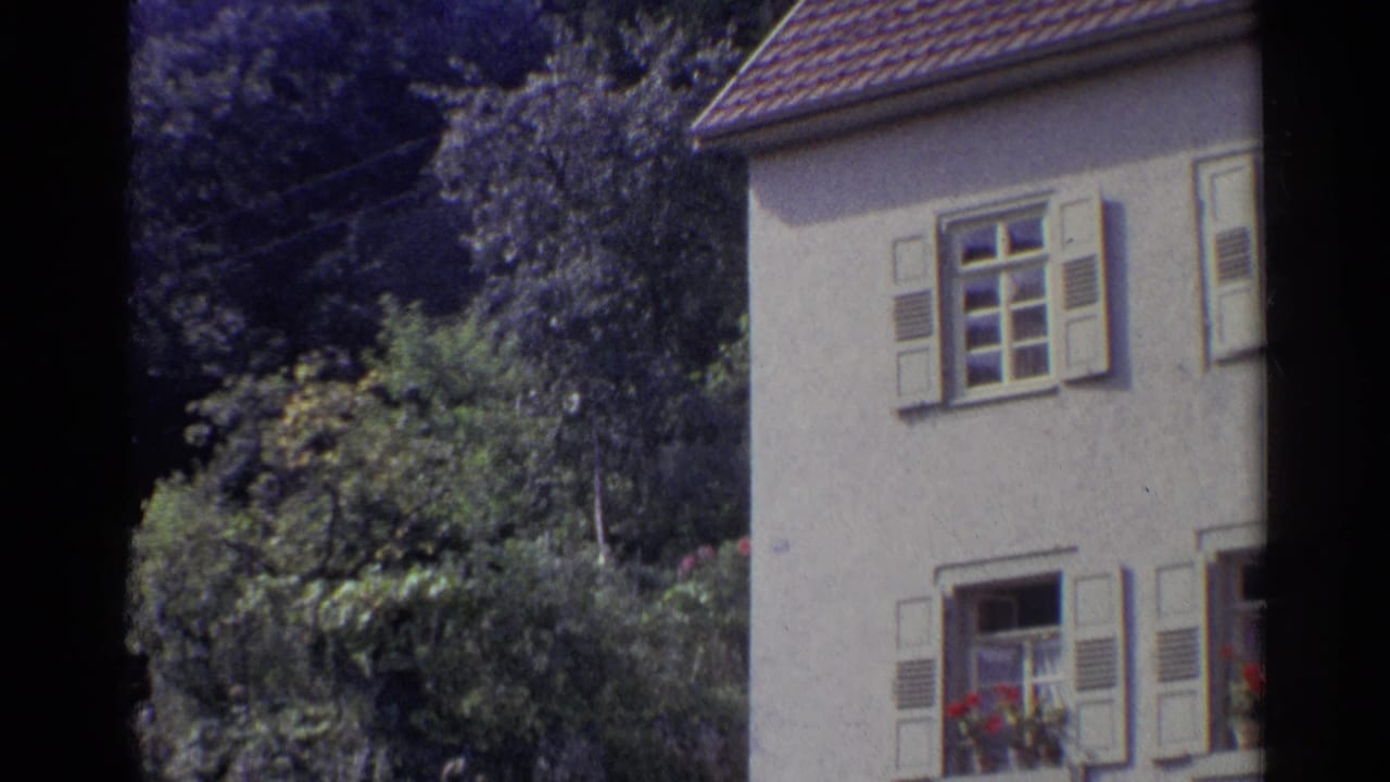 1961 German home-movie frame showing a pale house with shutters, flower boxes, trees, and thick garden foliage.