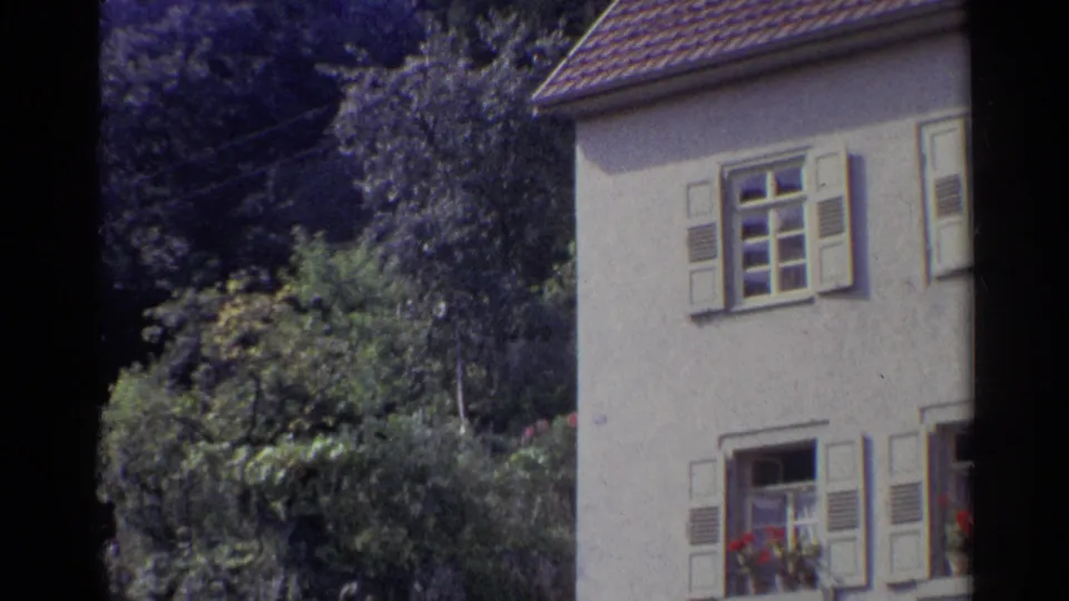 1961 German home-movie frame showing a pale house with shutters, flower boxes, trees, and thick garden foliage.
