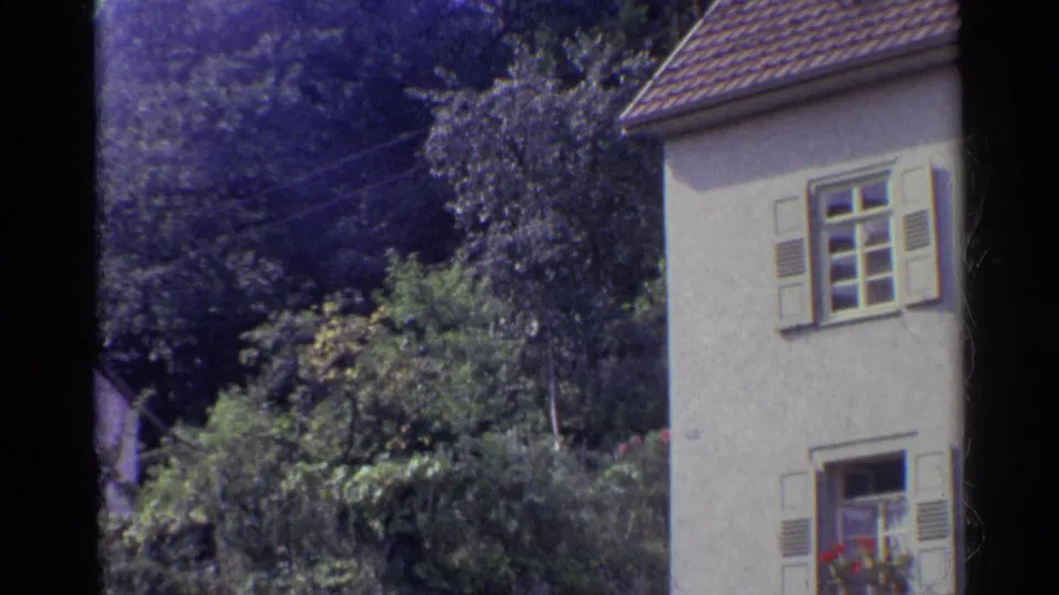1961 German residential exterior with shuttered windows, a tiled roof, flower boxes, and surrounding garden foliage.