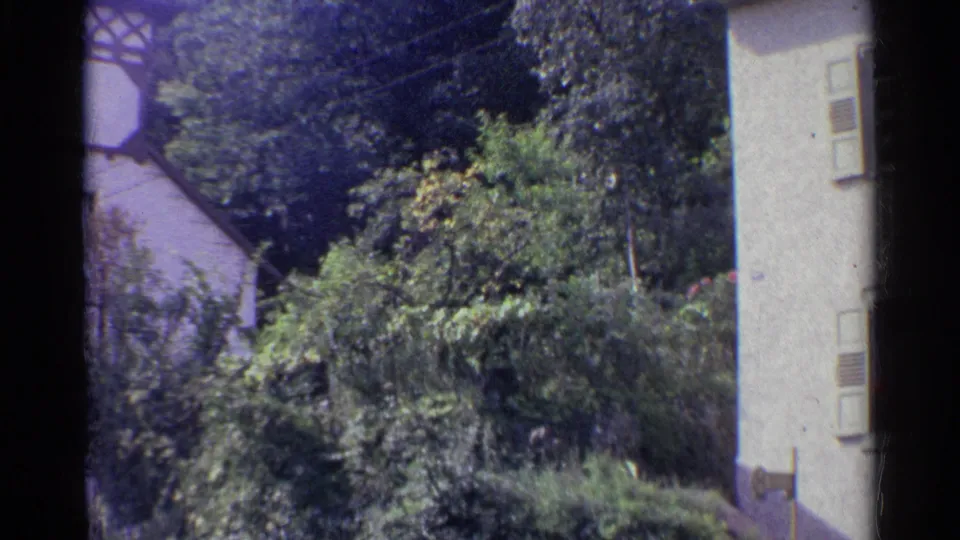 1961 Germany home-movie frame showing a pale residential building, leafy garden growth, and a street edge.