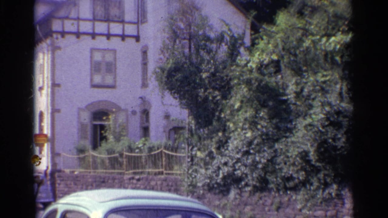 Home-movie view of a German residential facade with timber trim, leafy shrubs, stone wall, and the roof of an older car.