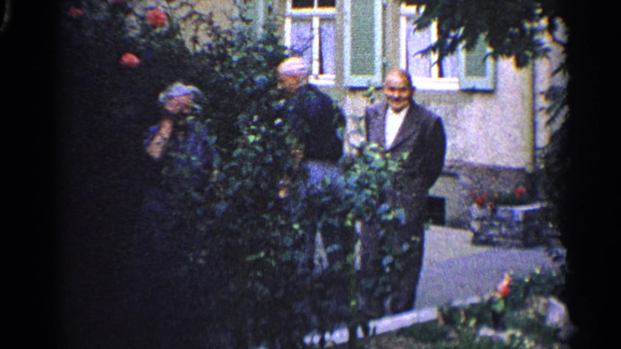 1961 home-movie frame showing adults in a garden beside a building, with foliage, flowers, windows, and a shaded walkway.
