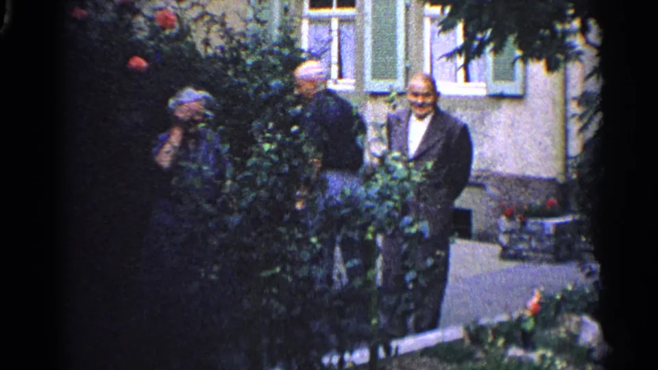 1961 home-movie frame showing adults in a garden beside a building, with foliage, flowers, windows, and a shaded walkway.