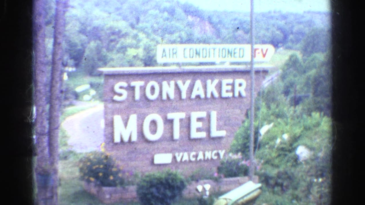1961 home-movie frame of a brick Stonyaker Motel sign marked air conditioned, TV, and vacancy, with trees, road, and hillside behind it.