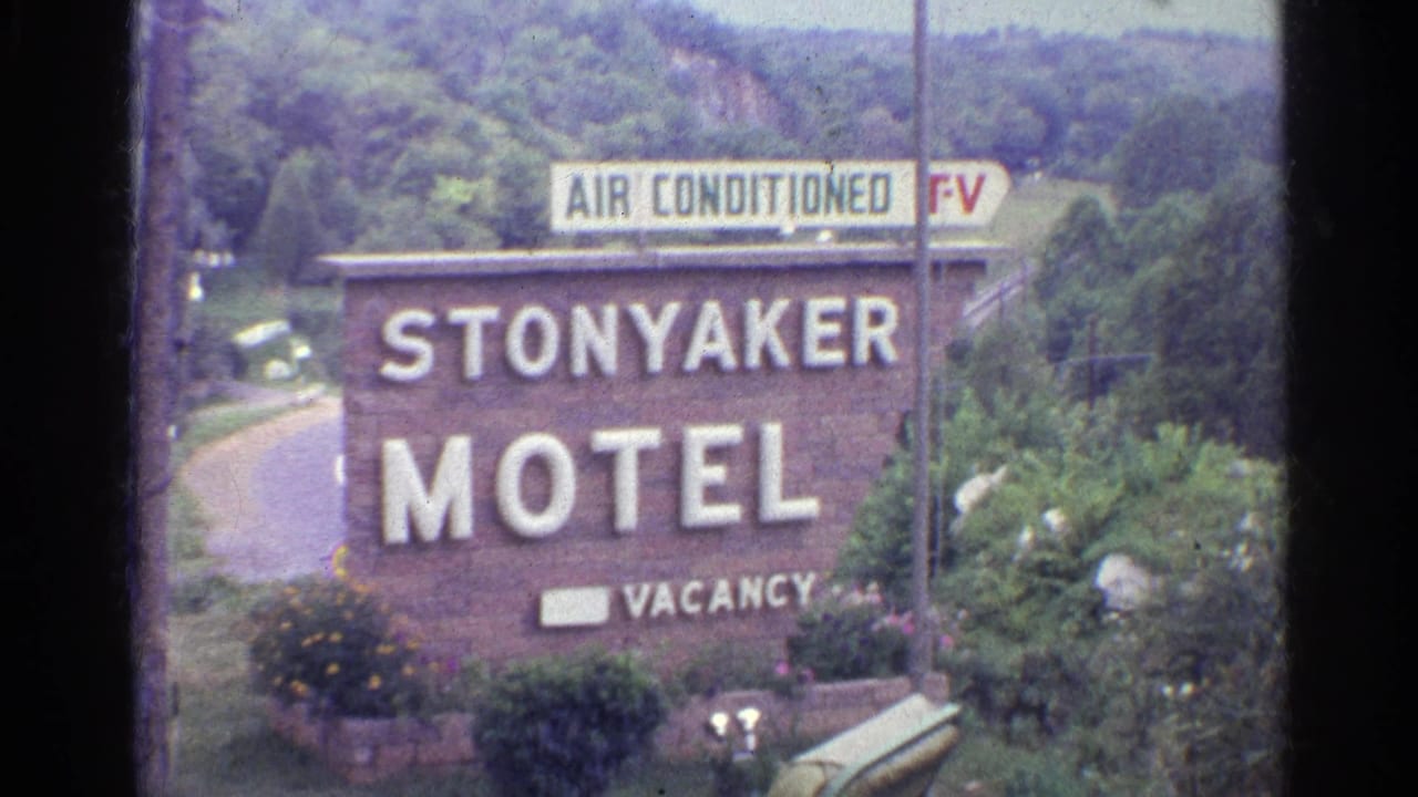 1961 home-movie frame of the Stonyaker Motel roadside sign with vacancy marker, flowers, shrubs, trees, and a pale sky beyond.