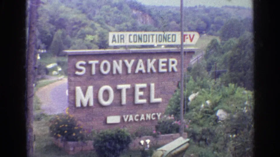 1961 home-movie frame of the Stonyaker Motel roadside sign with vacancy marker, flowers, shrubs, trees, and a pale sky beyond.