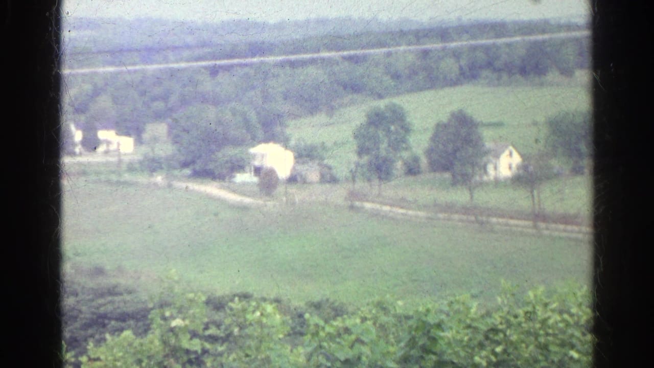 1961 Germany home-movie view of green fields, scattered trees, pale farmhouses, and a winding rural road in the distance.