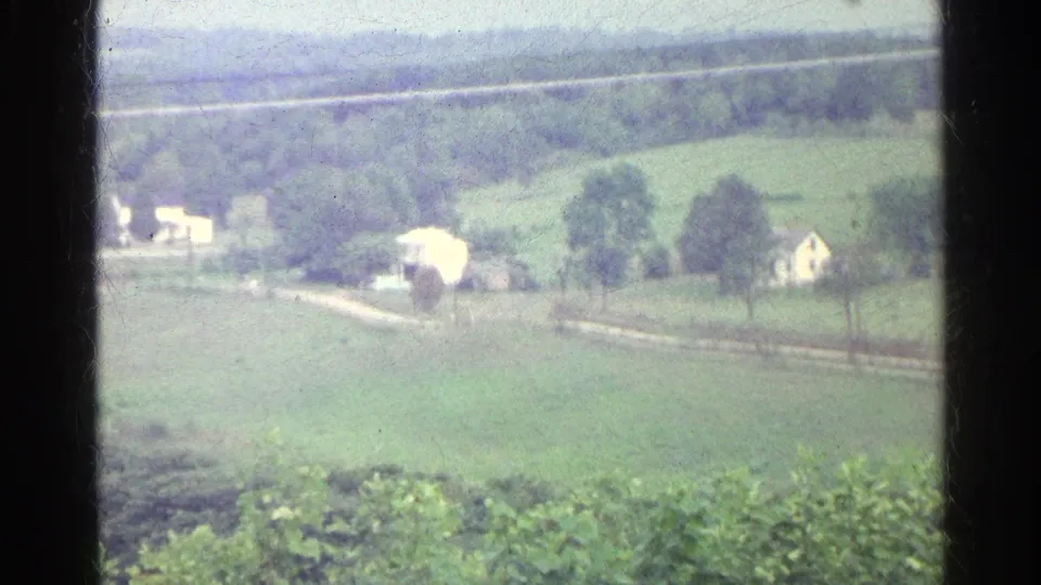 1961 Germany home-movie view of green fields, scattered trees, pale farmhouses, and a winding rural road in the distance.