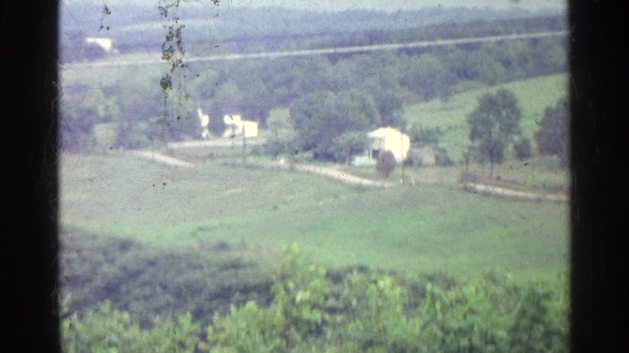 Rural valley with pale buildings, green fields, trees, and a winding road.