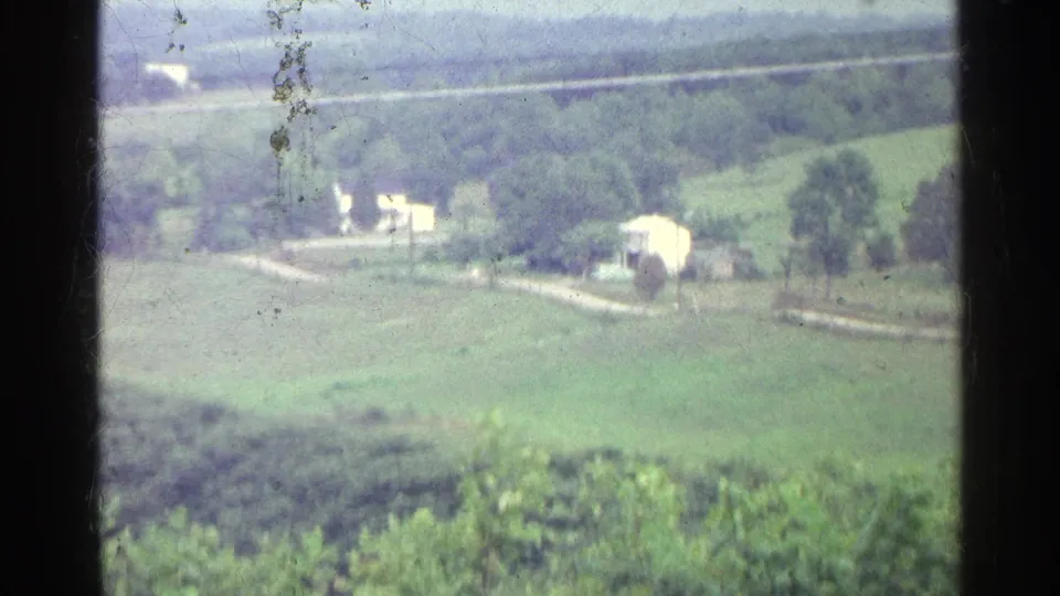 1961 Germany countryside with pale farm buildings, open green fields, tree lines, and a road curving through the landscape.