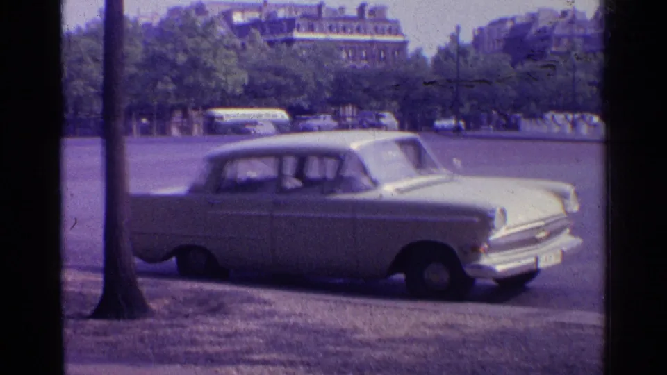 A white mid-century sedan remains parked at the curb in Paris, with a tree trunk, roadway, and background vehicles visible.