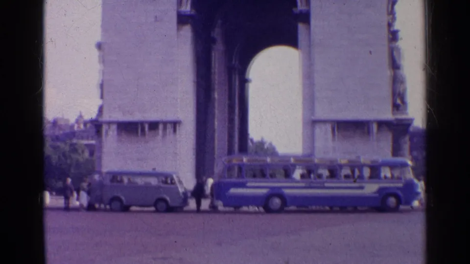 The Arc de Triomphe fills the background while a bus, small van, and pedestrians occupy the street-level foreground in Paris.