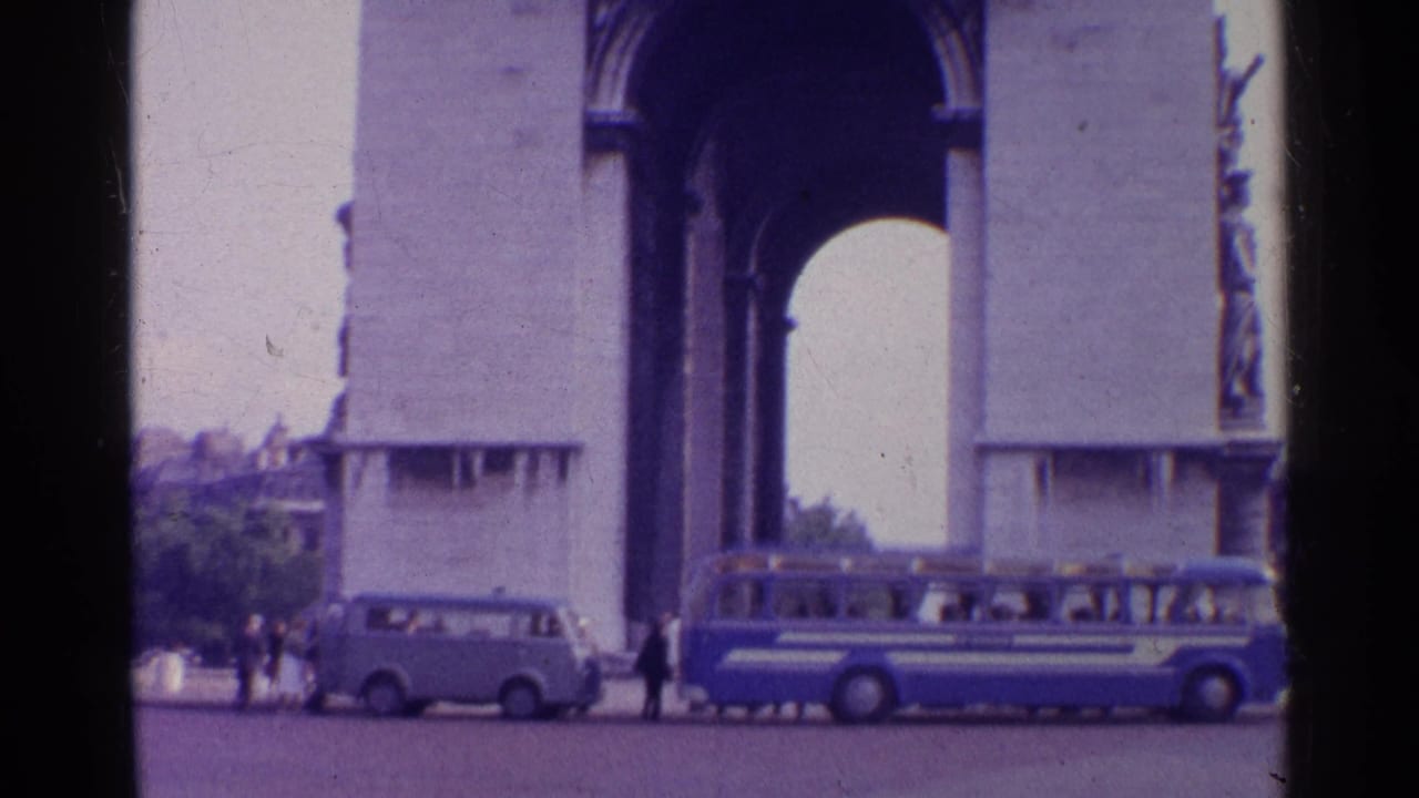 1961 8mm frame of the Arc de Triomphe in Paris with a blue-and-white bus, light van, and pedestrians near the base.