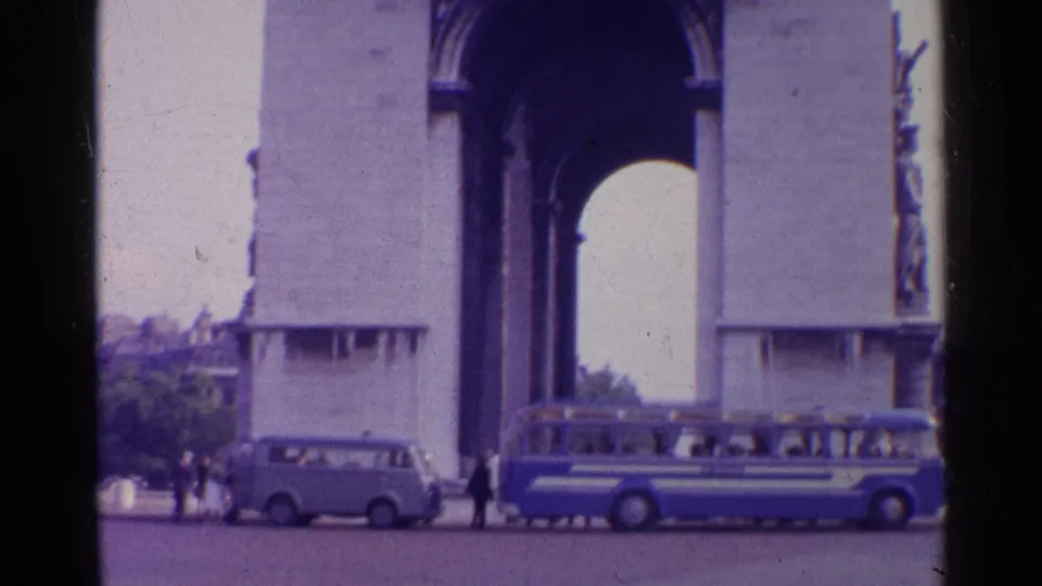 1961 8mm frame of the Arc de Triomphe in Paris with a blue-and-white bus, light van, and pedestrians near the base.