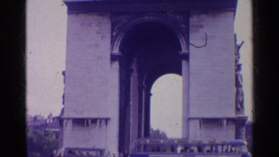 1961 Paris home-movie frame showing the Arc de Triomphe base as the camera begins to tilt upward from vehicles below.