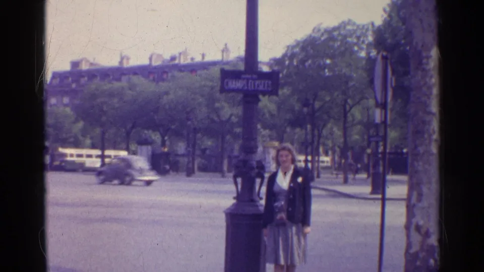 1961 Paris street scene with a woman posed beside a Champs-Elysees sign, passing cars, trees, and buildings in the background.
