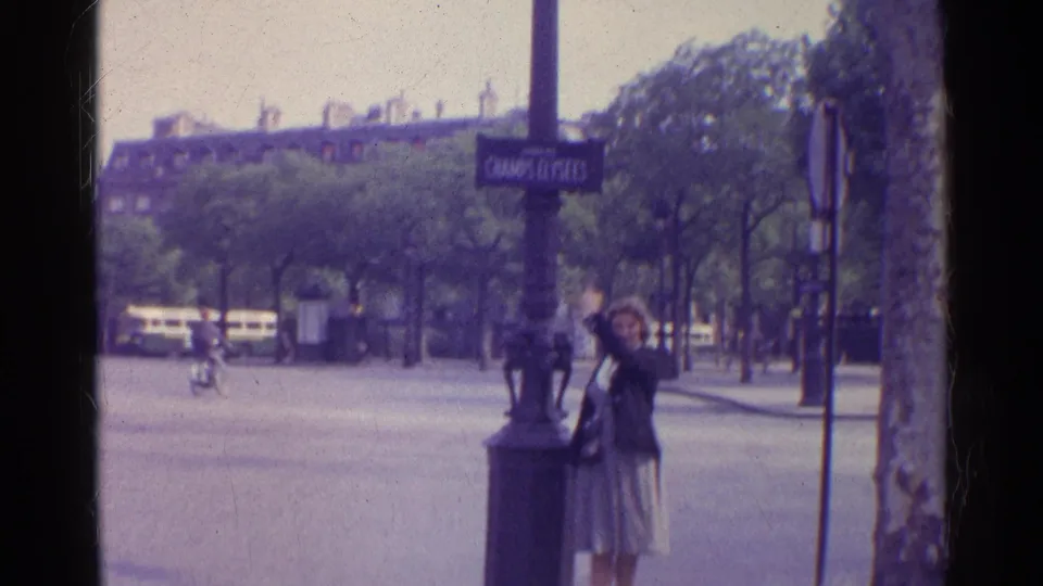 1961 Paris home-movie frame showing a woman posed near a Champs Elysees sign as cars pass along a tree-lined street.