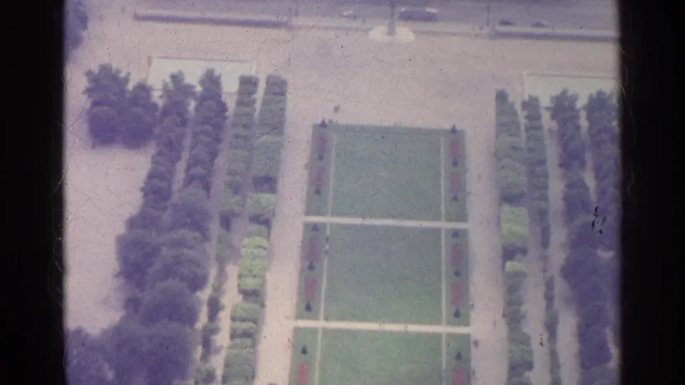 Paris 1961 8mm frame with a tilted overhead view of a rectangular lawn, pale walkways, hedges, trees, and nearby street activity.
