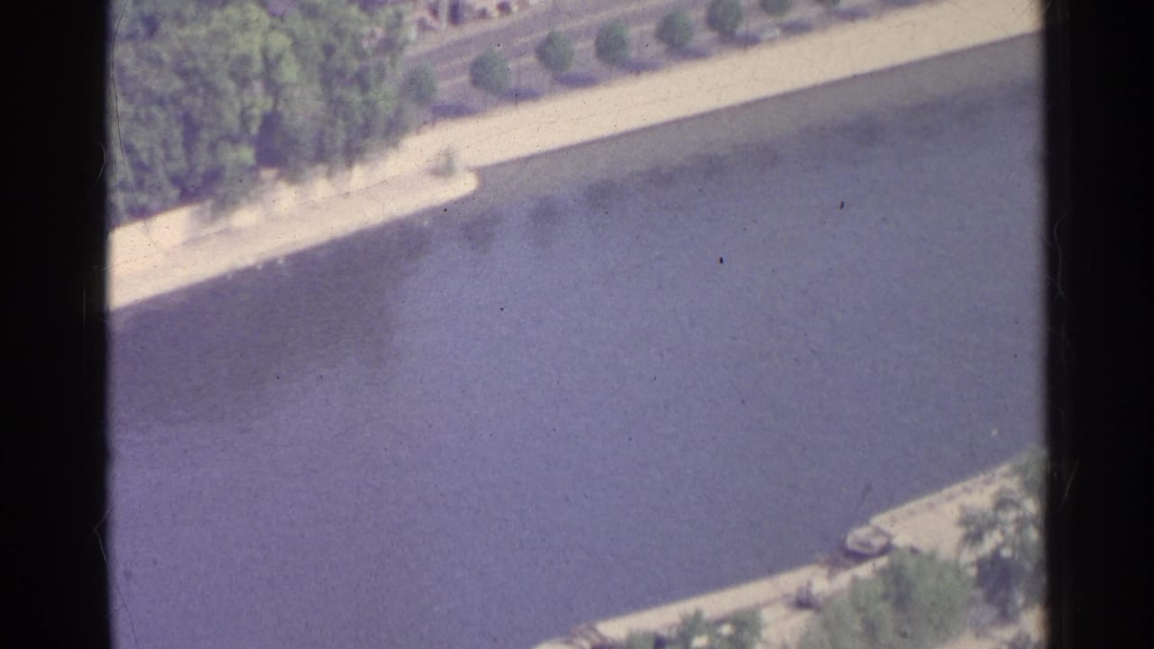 High-angle view of a river edge with trees, walkway, and part of a bridge or embankment.